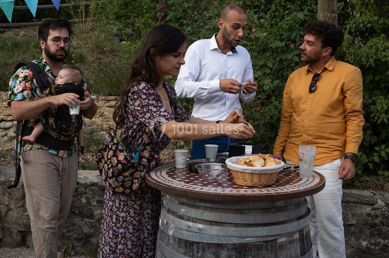 Des personnes discutent autour d'une table en tonneau, partageant des viennoiseries dans un cadre extérieur verdoyant.