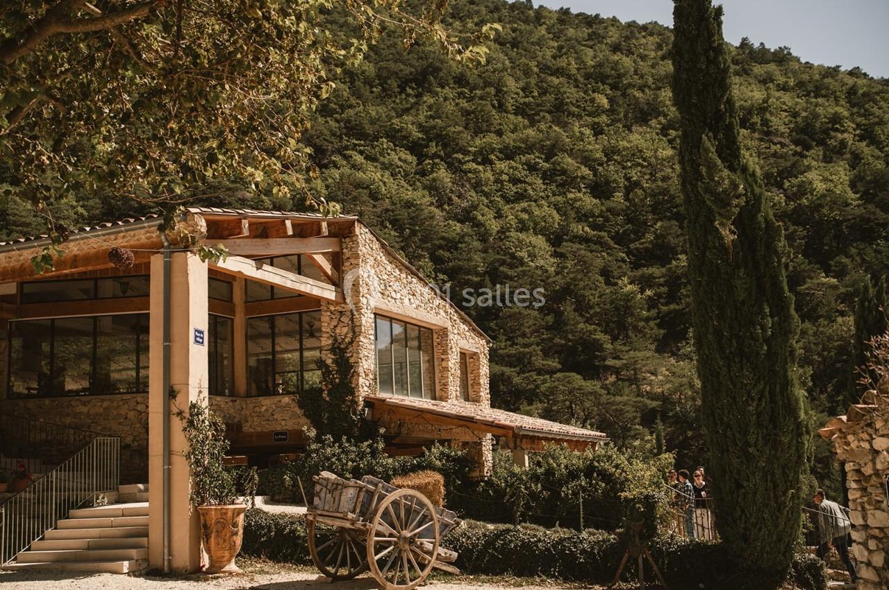 Bâtisse en pierre avec de grandes baies vitrées, entourée de végétation et d'un chariot en bois au premier plan.