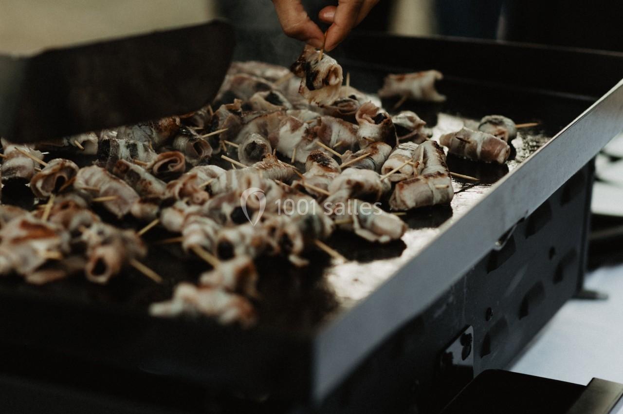 Une main retourne des brochettes de viande grillées sur une plancha noire lors d'une cuisson en extérieur.