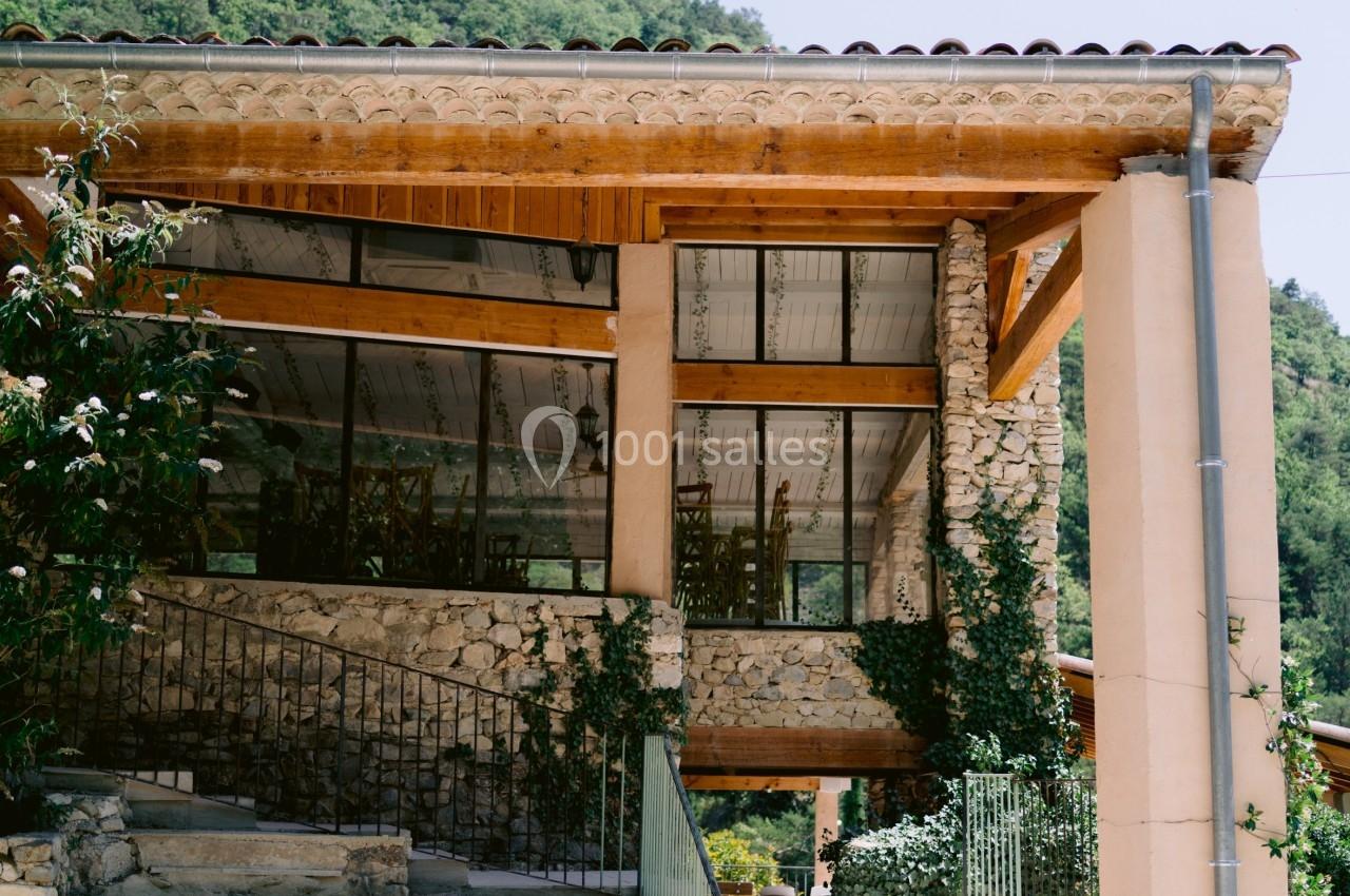 Façade d'un bâtiment en pierre et bois avec de grandes baies vitrées, entouré de végétation et d'une colline verdoyante.