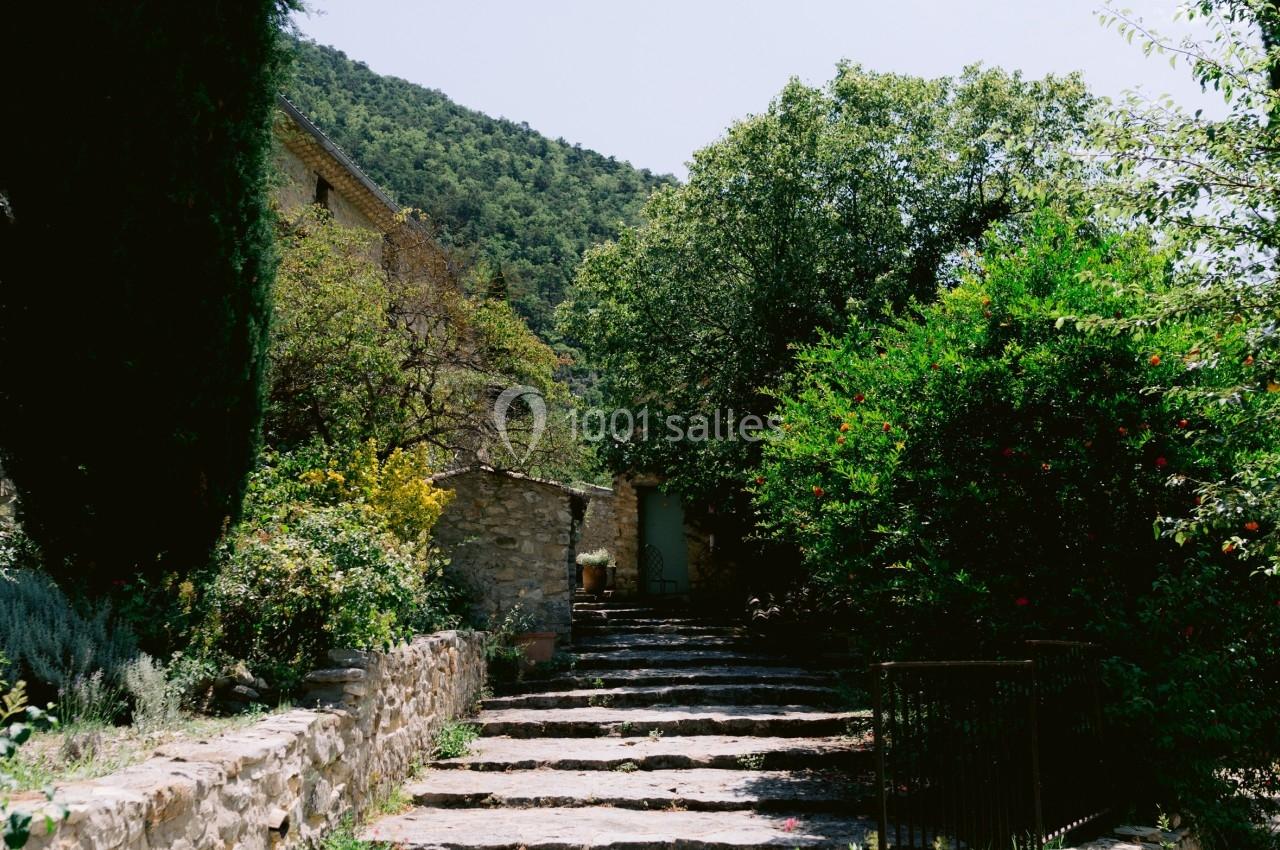 Escalier en pierre bordé de végétation, menant à une maison en pierre dans un cadre naturel ensoleillé.