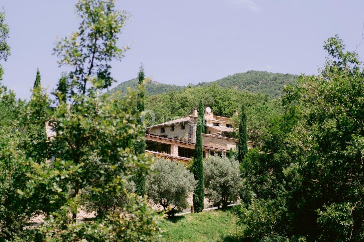Maison en pierre entourée de cyprès et de végétation, située dans un paysage vallonné sous un ciel dégagé.