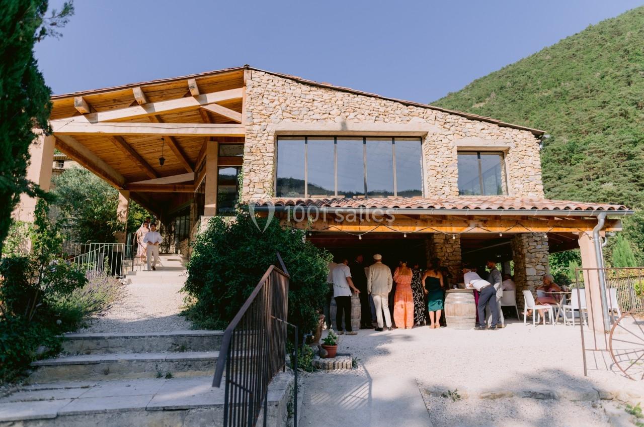 Façade d'un bâtiment en pierre avec une terrasse couverte, des invités rassemblés à l'extérieur par une journée ensoleillée.
