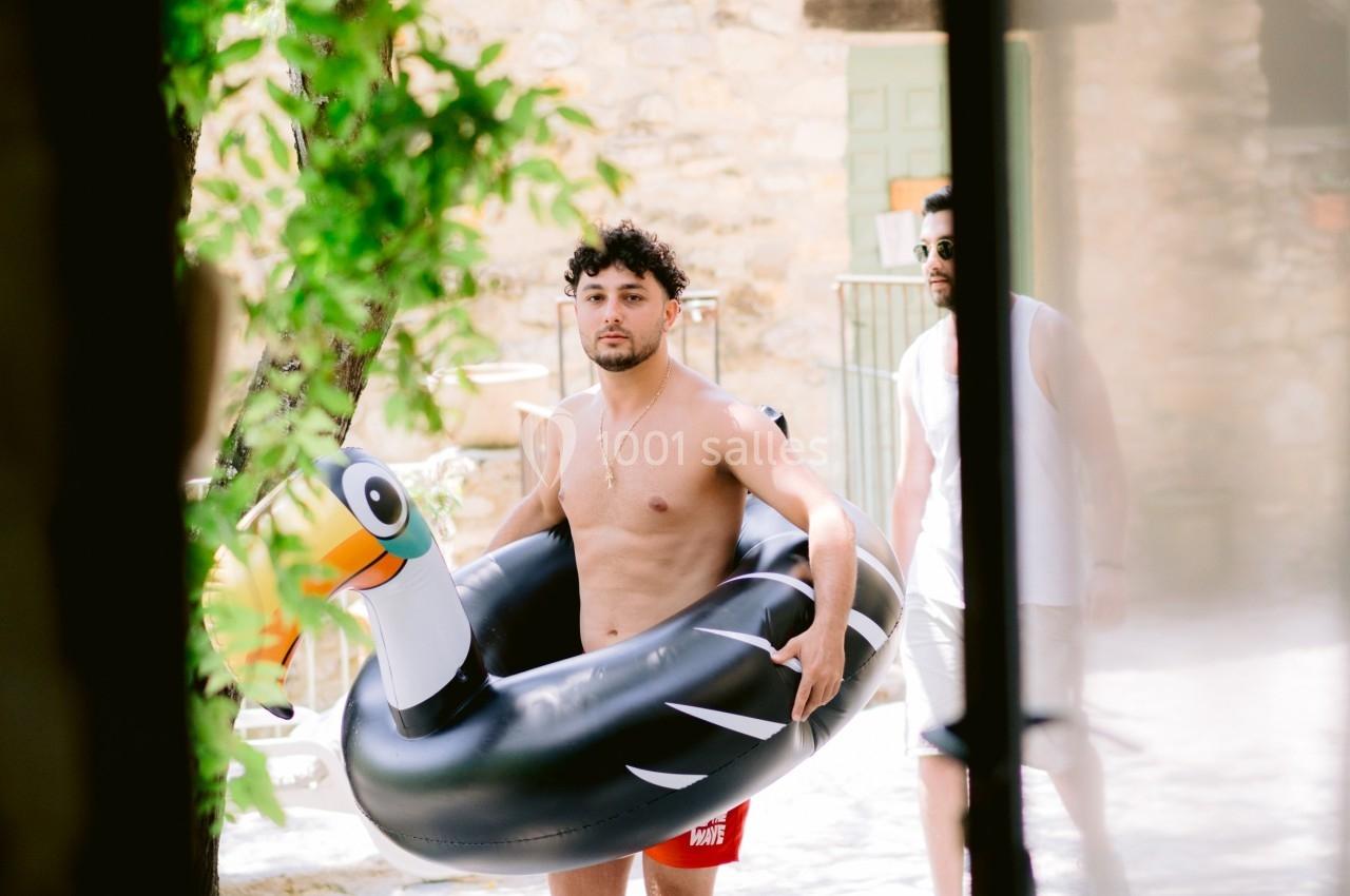 Un homme torse nu portant une bouée noire en forme d'oiseau marche dans une cour ensoleillée.