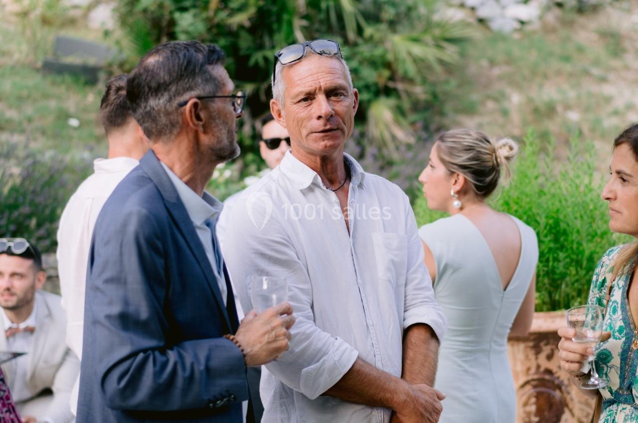 Un groupe de personnes discute dans un jardin, certaines tenant des verres, avec un plat en terre cuite au premier plan.