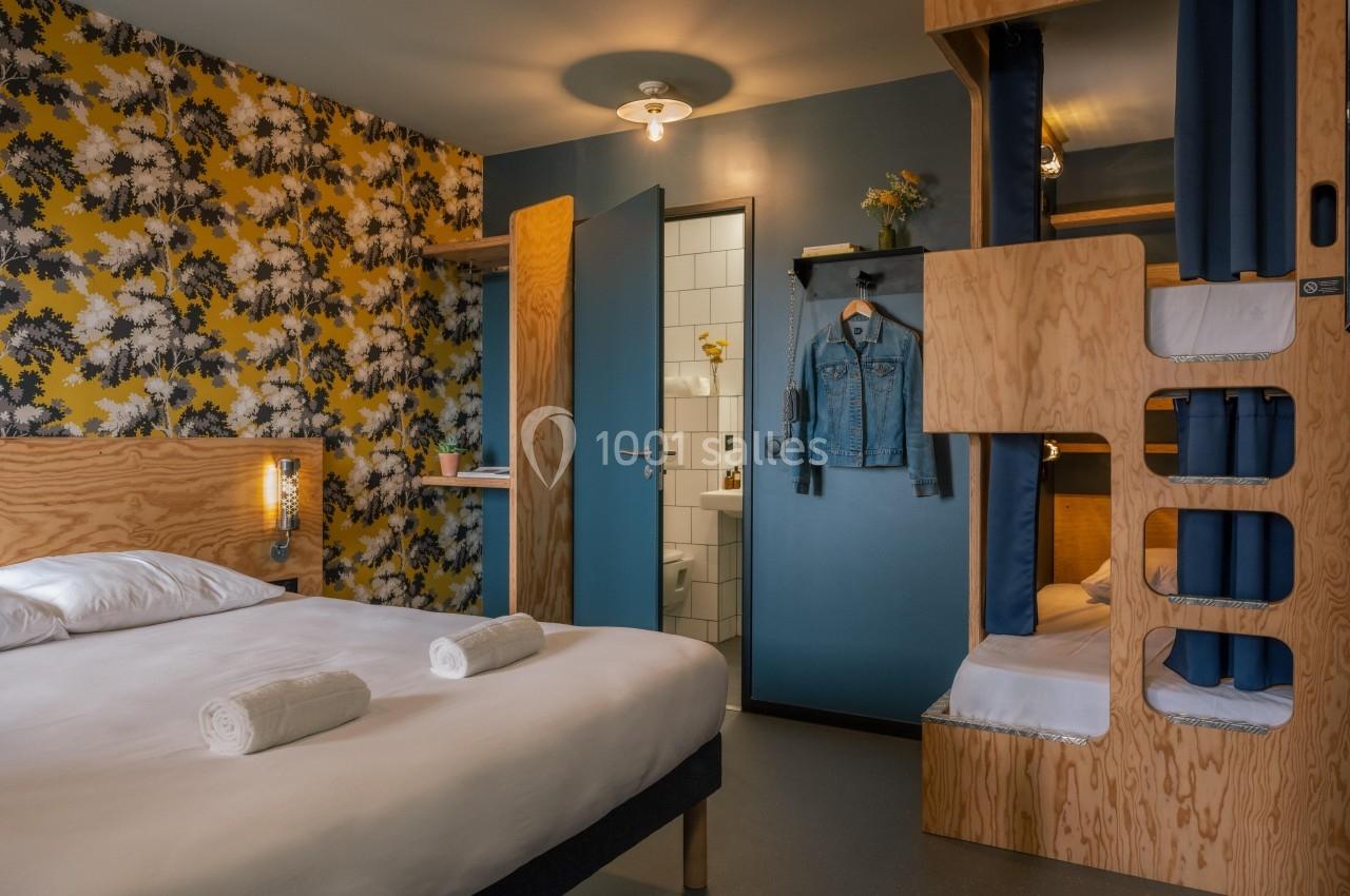 Chambre avec lit double, lits superposés en bois, mur fleuri, salle de bain ouverte et éclairage chaleureux.