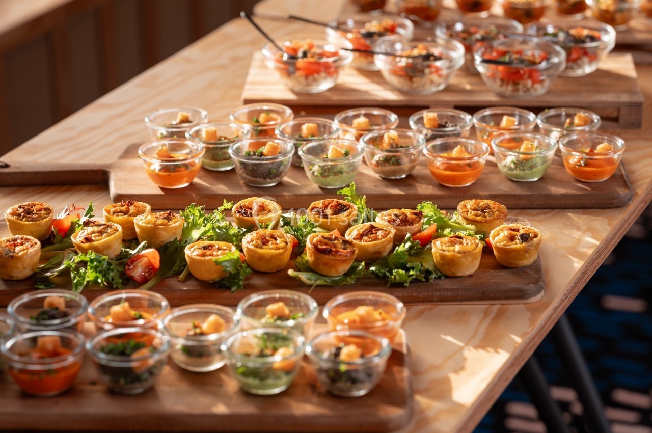 Plateaux en bois garnis de verrines colorées et de mini-quiches, disposés sur une table ensoleillée.