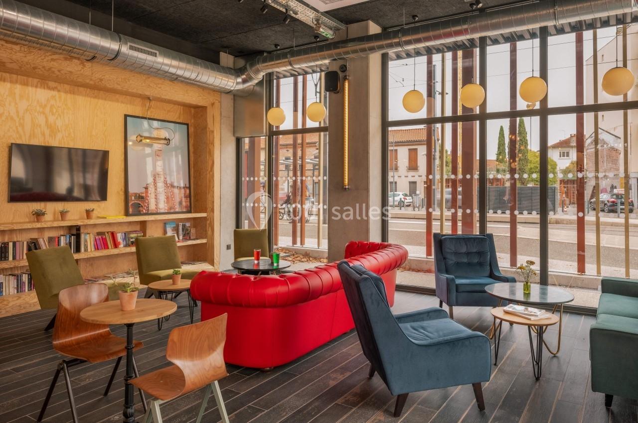 Espace lounge avec canapé rouge, fauteuils bleus, tables en bois et grande baie vitrée donnant sur l'extérieur.