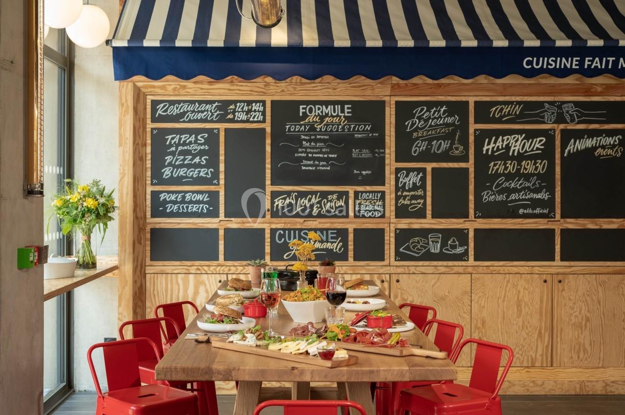 Table en bois garnie de plats variés, entourée de chaises rouges, devant un mur avec menus écrits à la craie.