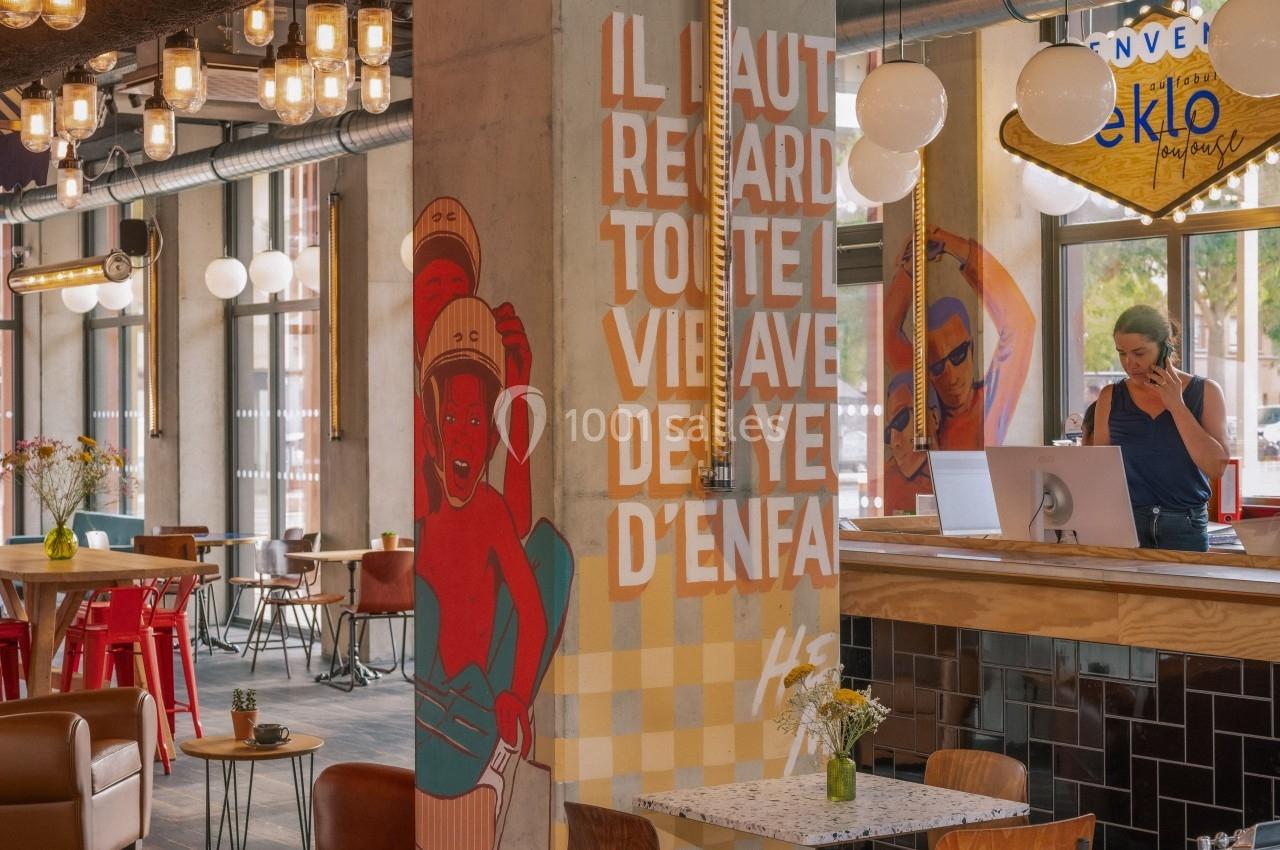 Intérieur d'un café moderne avec une fresque colorée, des tables, des chaises et une personne au comptoir.