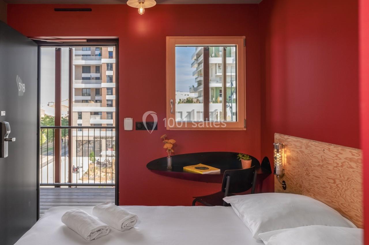 Chambre avec mur rouge, lit double, bureau noir, fenêtre et porte donnant sur un balcon avec vue sur des immeubles modernes.