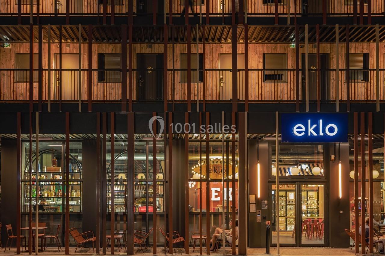 Façade d'un bâtiment moderne avec enseigne lumineuse ’eklo’, terrasse aménagée et intérieur éclairé visible.