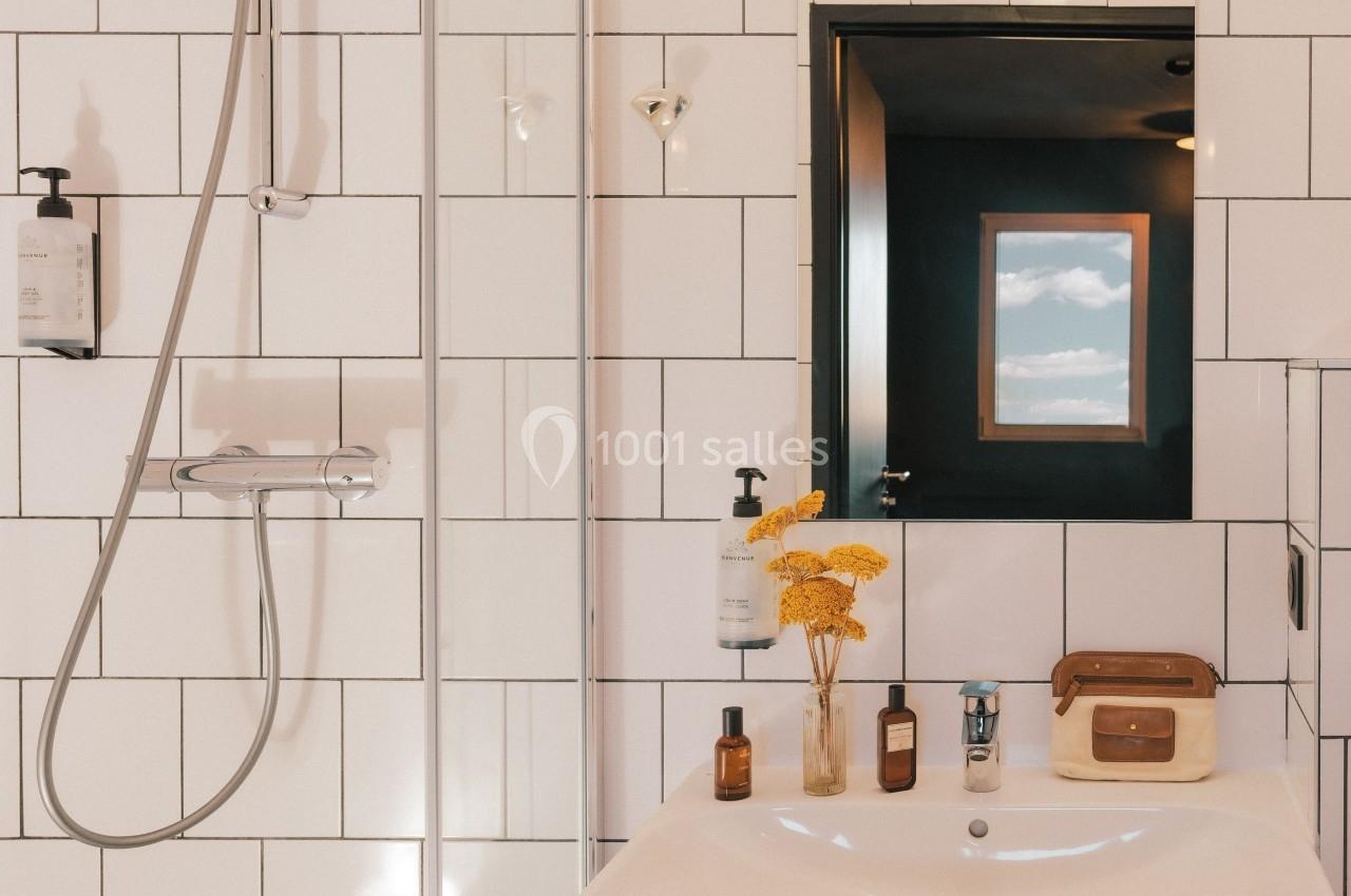 Salle de bain avec douche, lavabo, miroir, flacons et fleurs jaunes, carrelage blanc et fenêtre en arrière-plan.