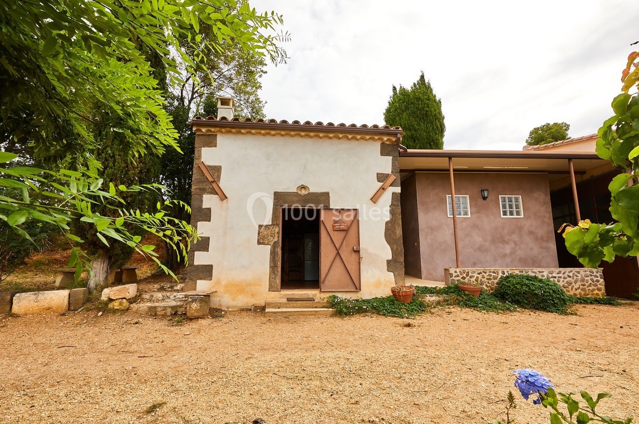 Petite maison en pierre avec porte en bois, entourée de végétation et située sur un terrain en gravier.