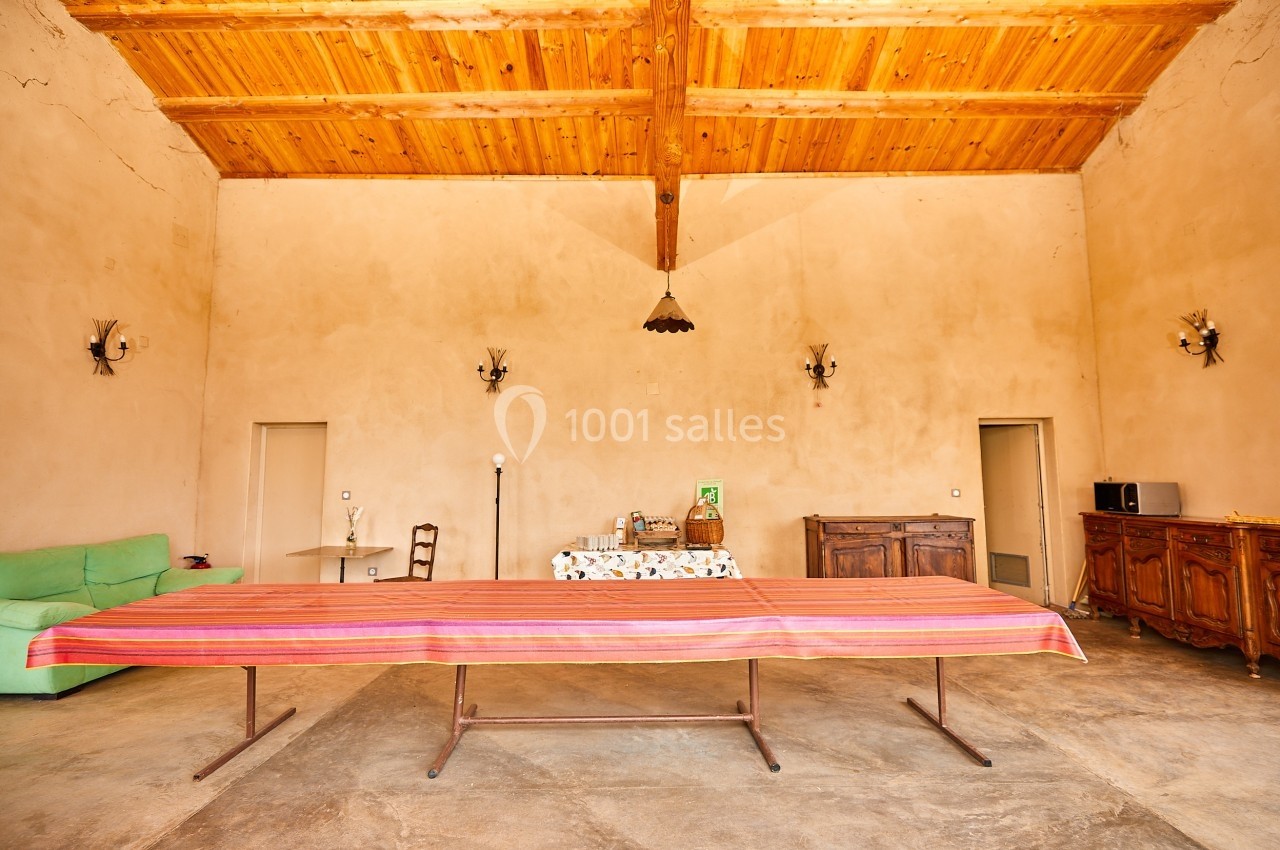 Salle avec plafond en bois, grande table recouverte d'une nappe colorée, canapé vert et meubles anciens en arrière-plan.