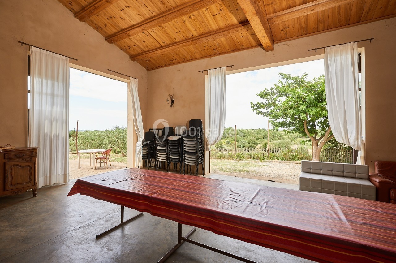 Salle lumineuse avec table recouverte d'un tissu coloré, chaises empilées, grandes ouvertures sur un jardin.