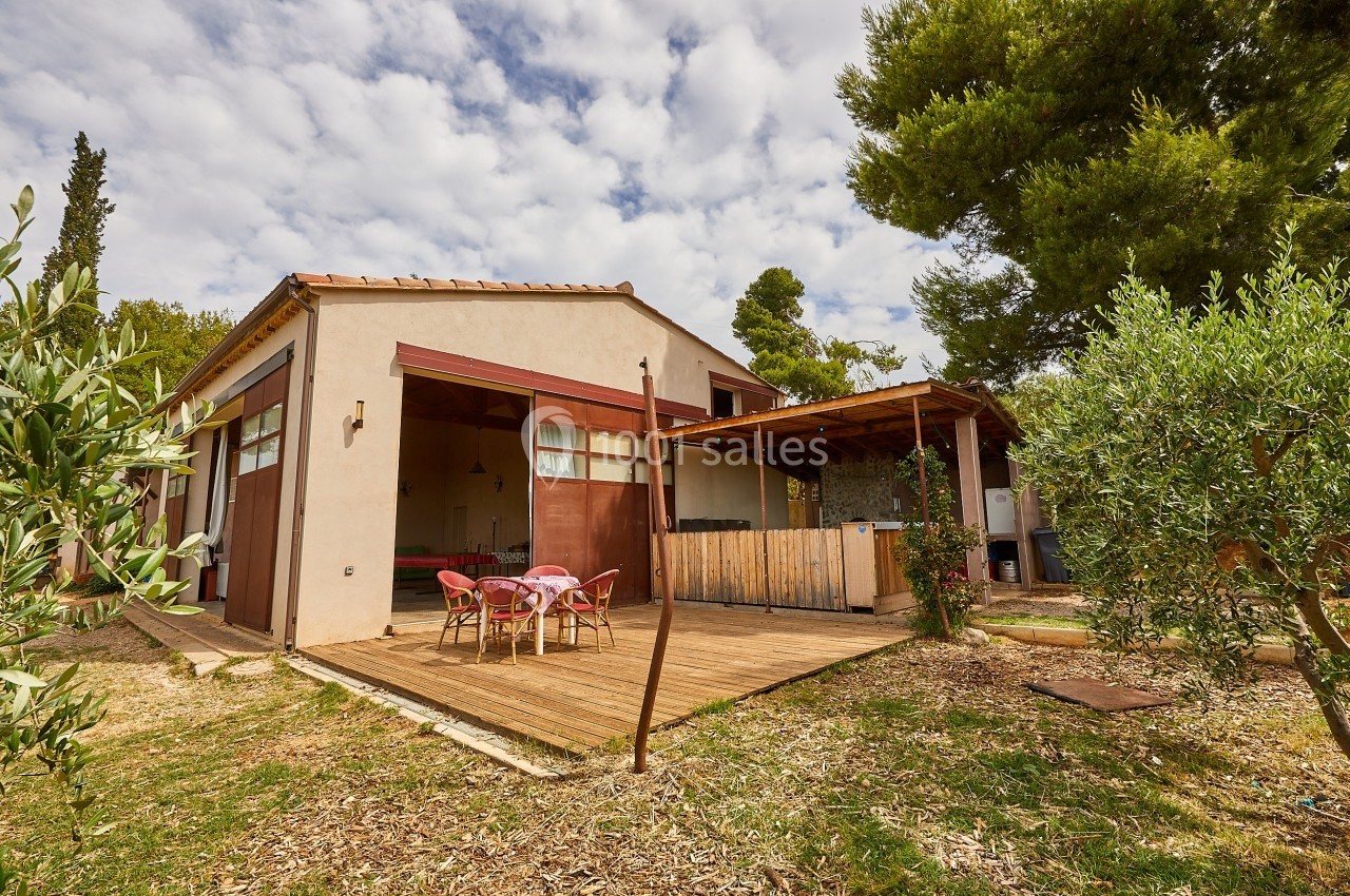 Maison avec terrasse en bois, table dressée, pergola et arbres environnants sous un ciel partiellement nuageux.