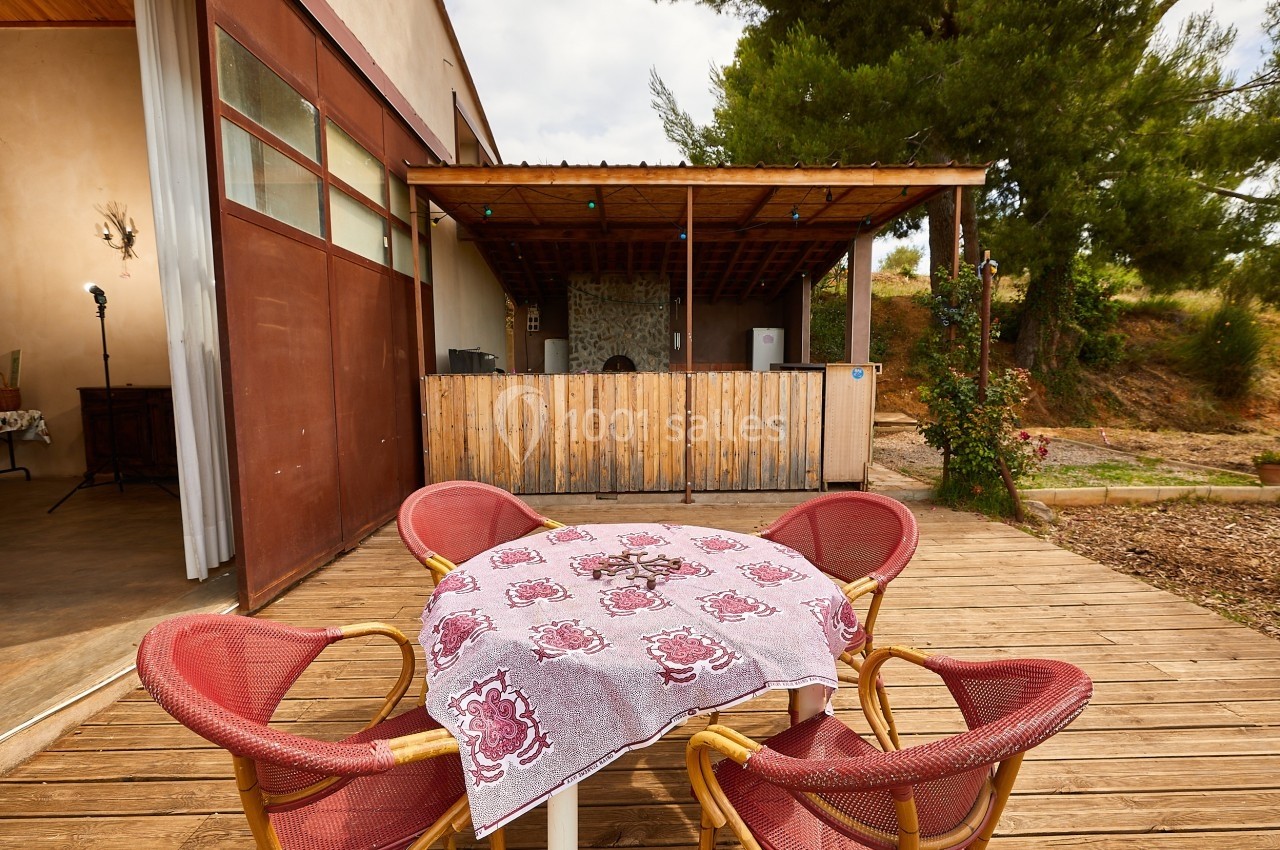 Table en plein air avec nappe à motifs, entourée de chaises rouges, sur une terrasse en bois près d'un espace couvert.