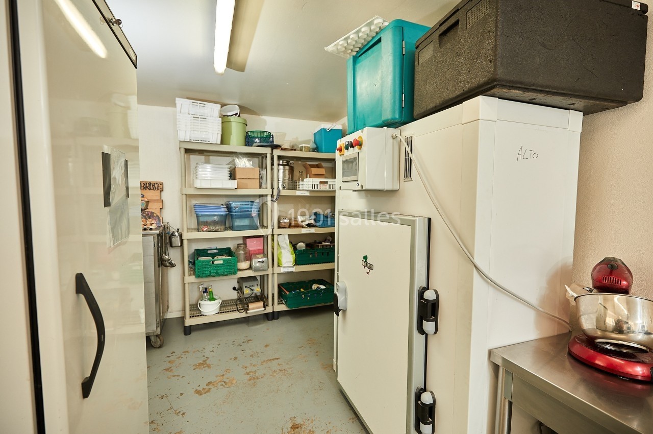 Salle de stockage avec étagères remplies de boîtes et d'ustensiles, réfrigérateur industriel et plan de travail en métal.