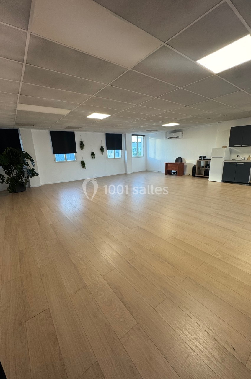 Salle lumineuse avec parquet clair, murs blancs, plantes décoratives, fenêtres et coin kitchenette équipé.