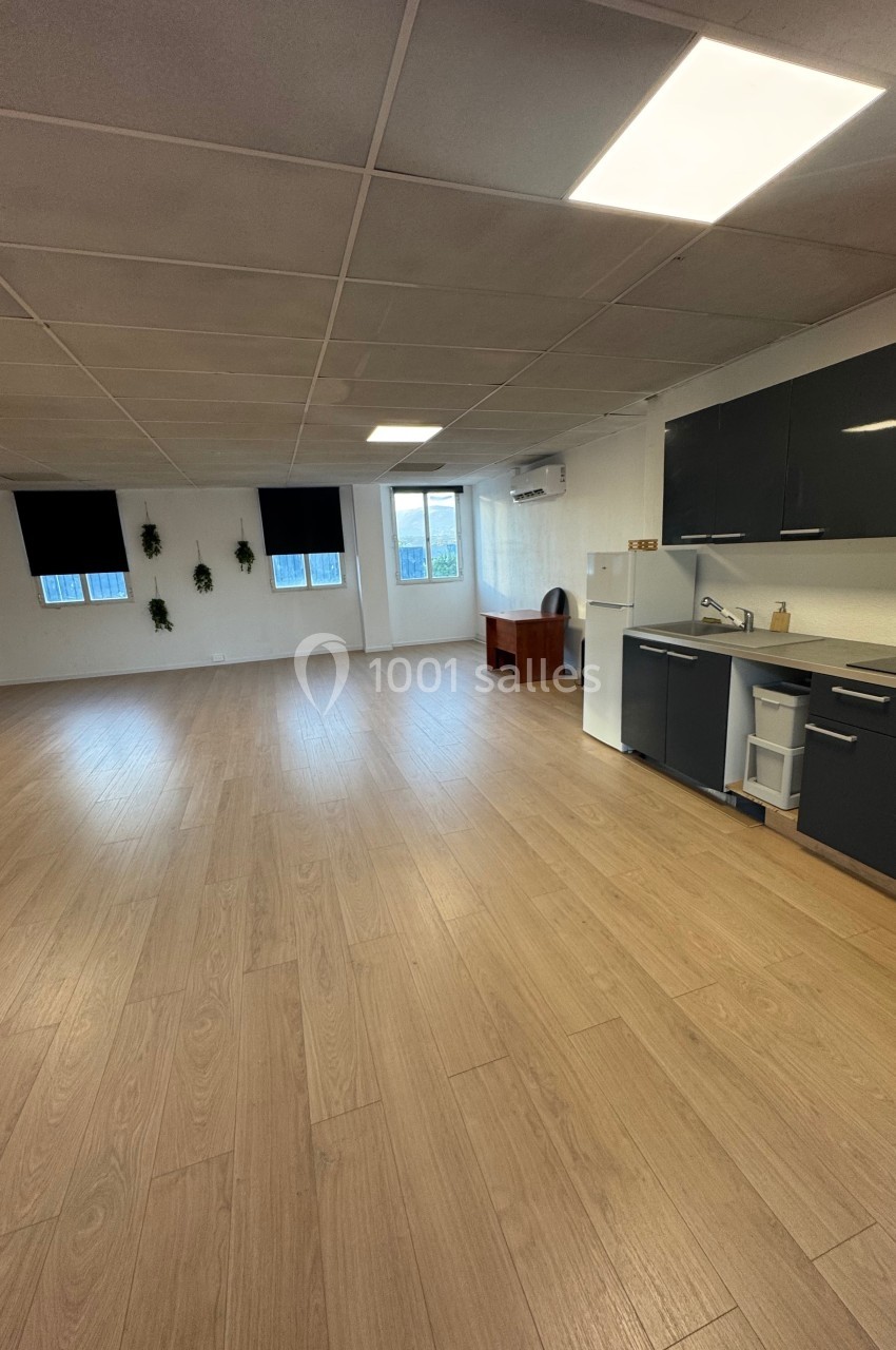 Salle lumineuse avec parquet, kitchenette équipée, bureau en bois, fenêtres et murs blancs décorés de plantes.