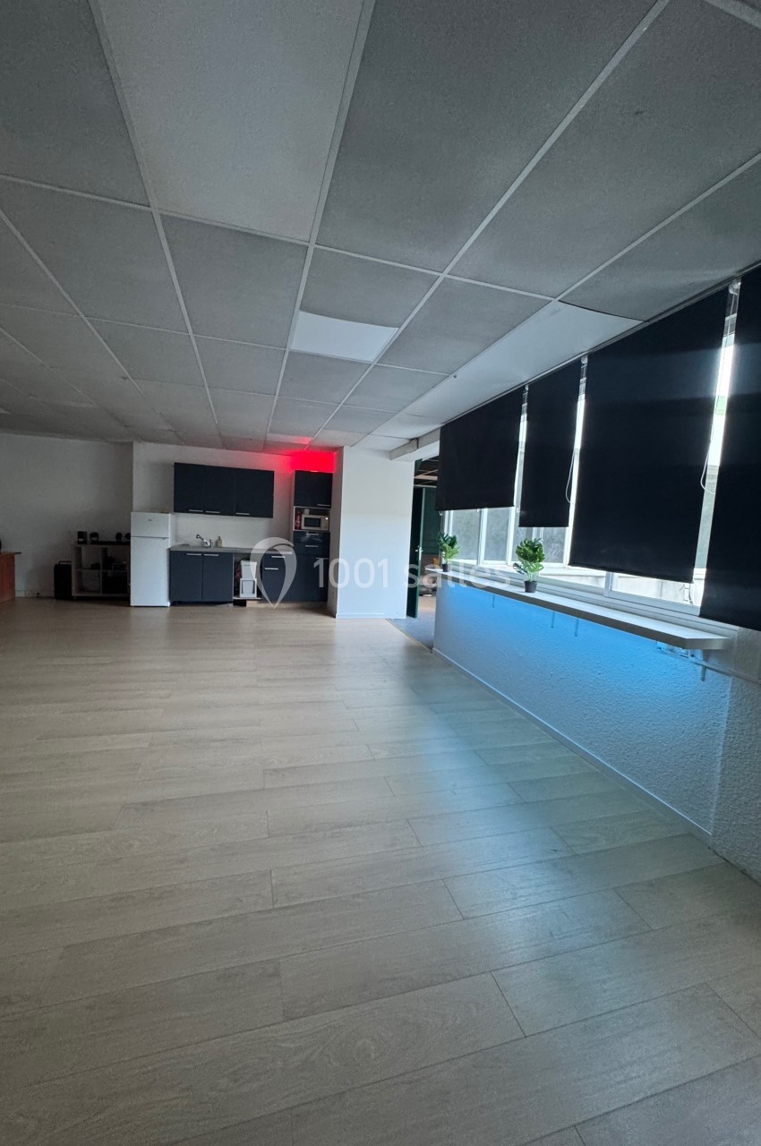 Salle lumineuse avec parquet clair, cuisine équipée au fond, stores noirs partiellement baissés sur les fenêtres.