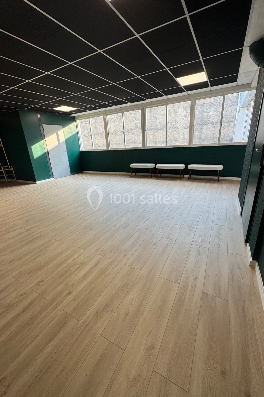 Salle vide avec parquet clair, murs verts, plafond noir et grande baie vitrée laissant entrer la lumière naturelle.