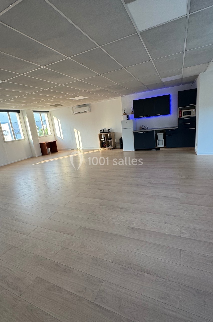 Salle lumineuse avec parquet clair, cuisine équipée au fond, murs blancs et grandes fenêtres laissant entrer la lumière…