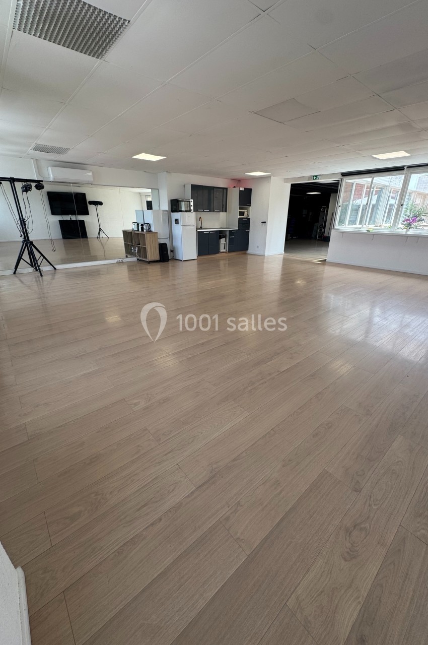 Salle lumineuse avec parquet en bois clair, équipée d'une cuisine ouverte, de miroirs et de matériel audiovisuel.