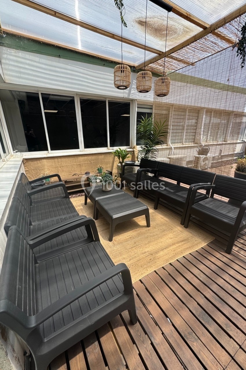 Terrasse aménagée avec des bancs noirs, une table basse, un tapis beige et des suspensions en rotin sous une pergola.