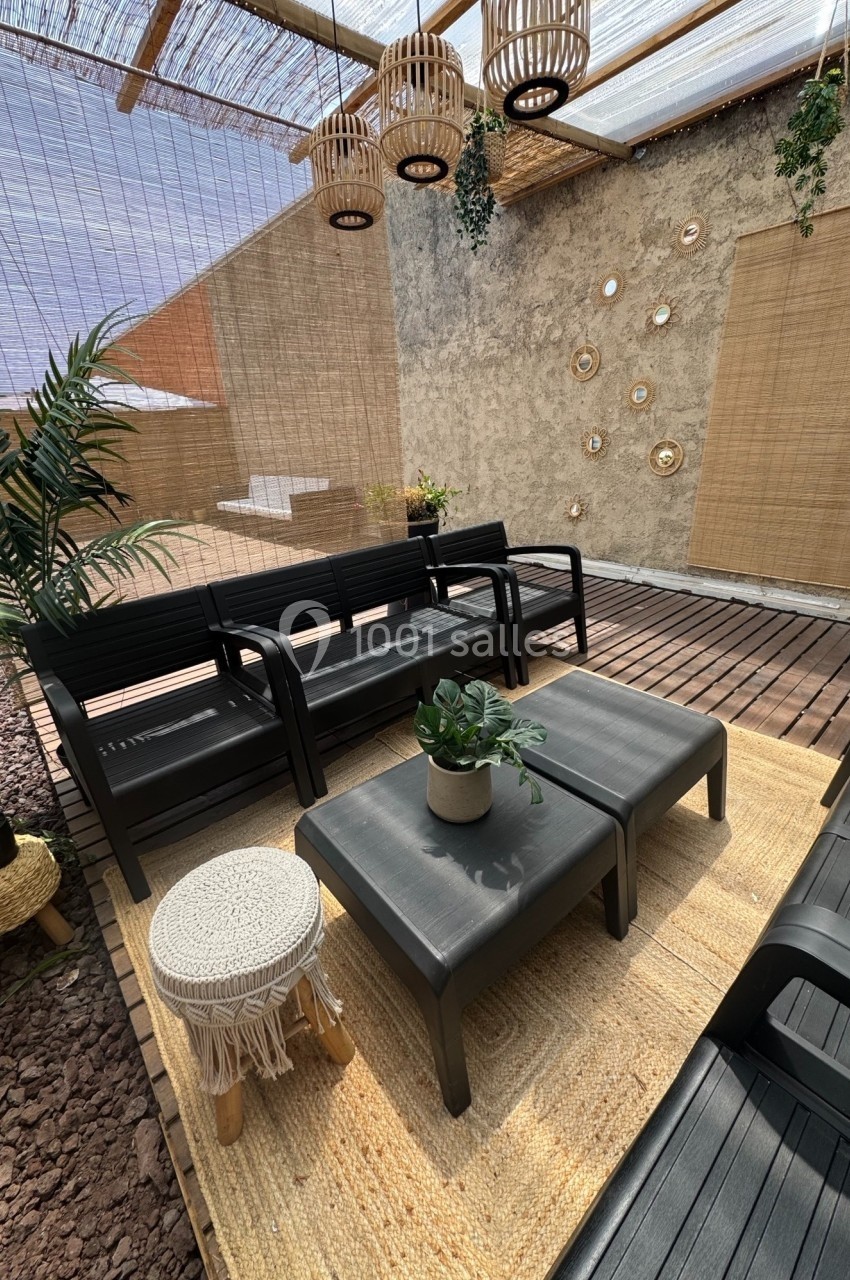 Salon extérieur avec mobilier en plastique noir, tapis en fibres naturelles, plantes et luminaires suspendus en rotin.