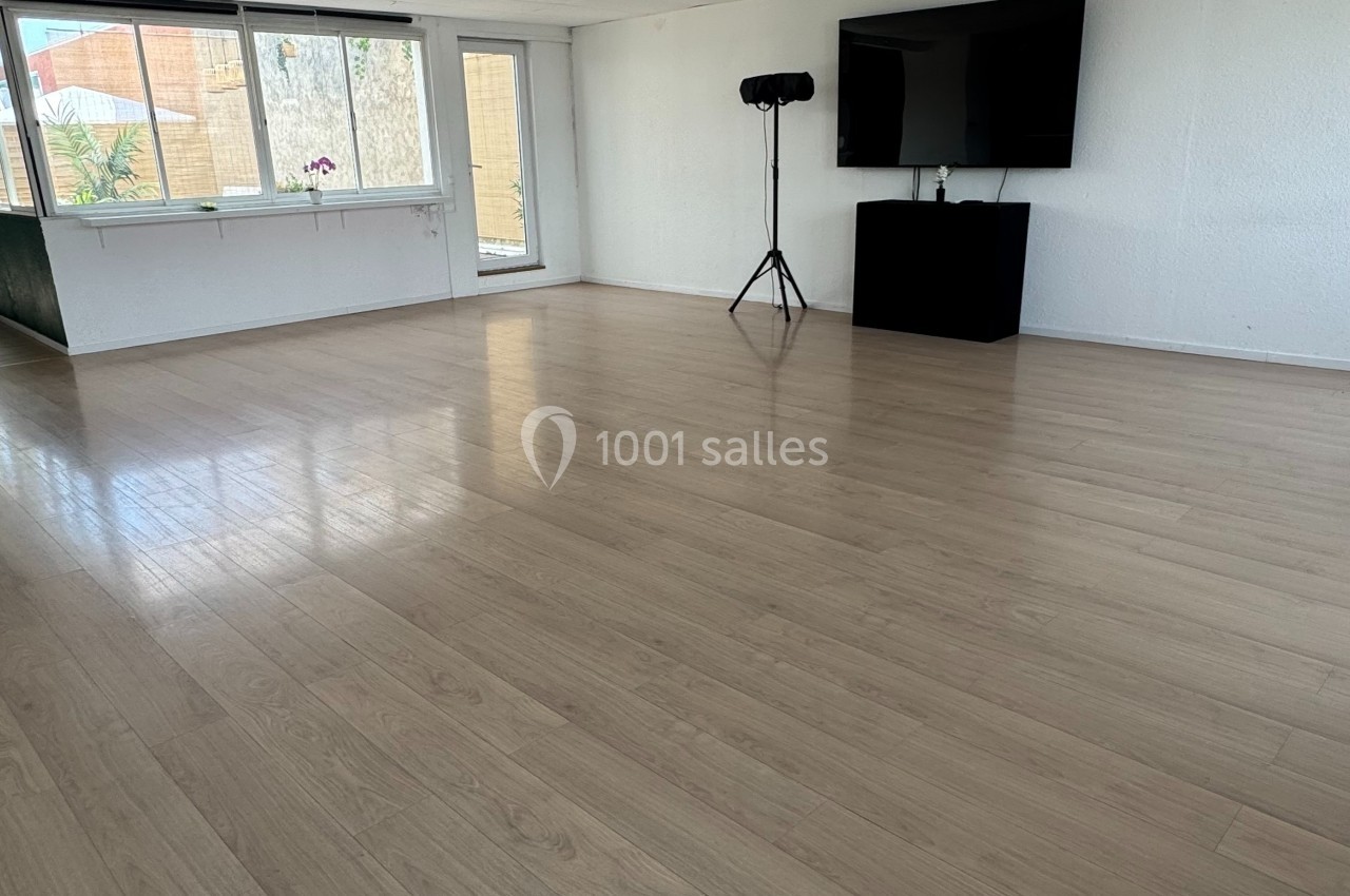 Salle lumineuse avec parquet clair, grande fenêtre, télévision murale et trépied dans un coin.