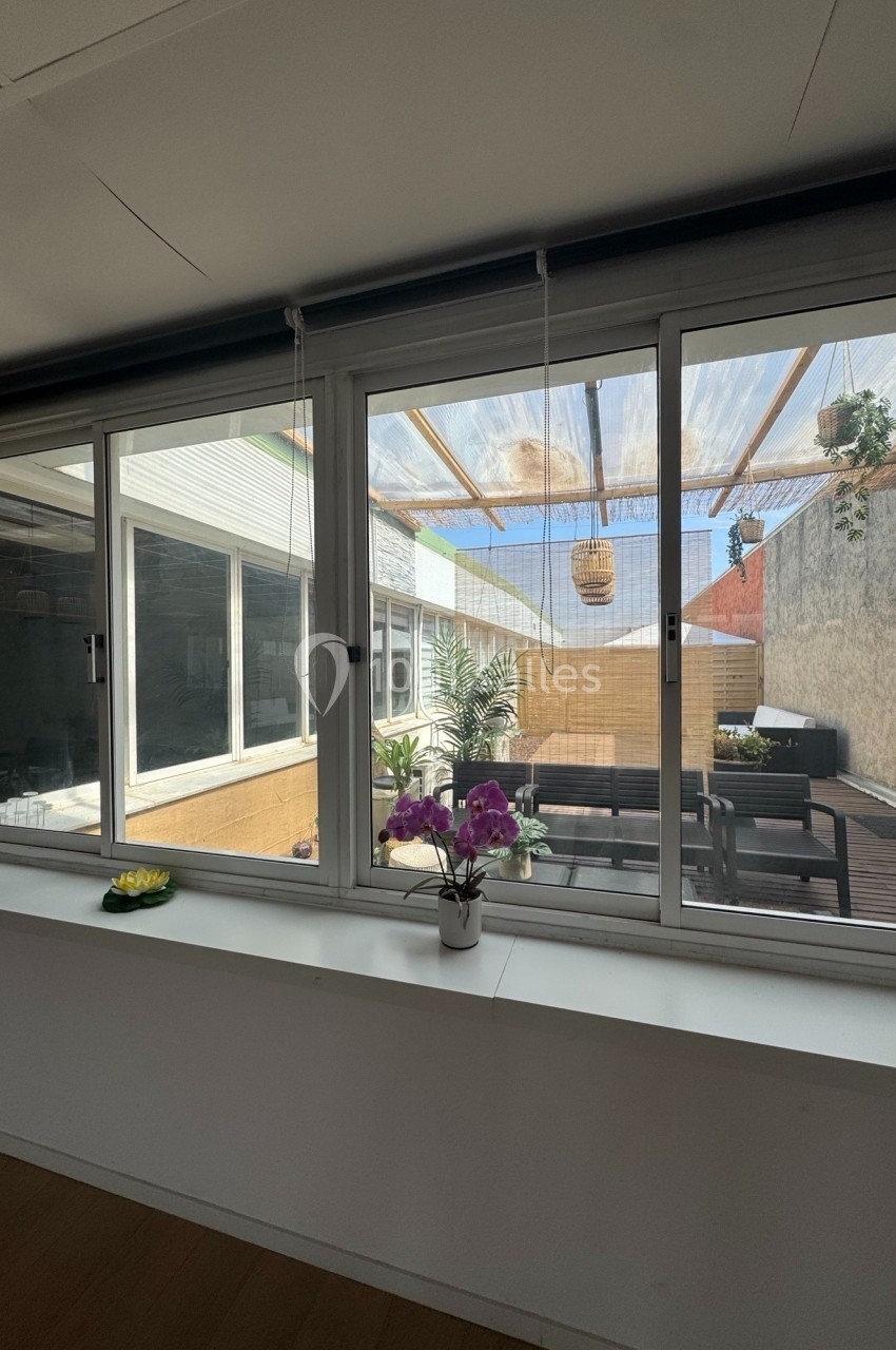 Vue depuis une fenêtre sur une terrasse ensoleillée avec table, plantes et toit en pergola translucide.
