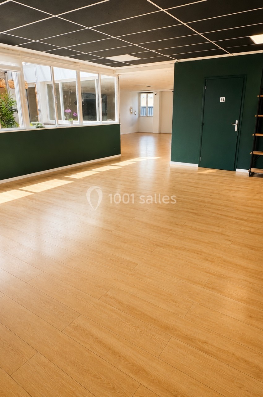 Salle lumineuse avec parquet clair, murs verts et blancs, grandes fenêtres et étagère dans un coin.