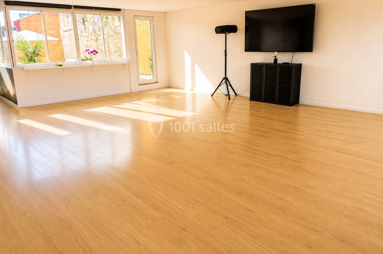 Salle lumineuse avec parquet en bois clair, grande fenêtre, télévision murale et trépied dans un coin.