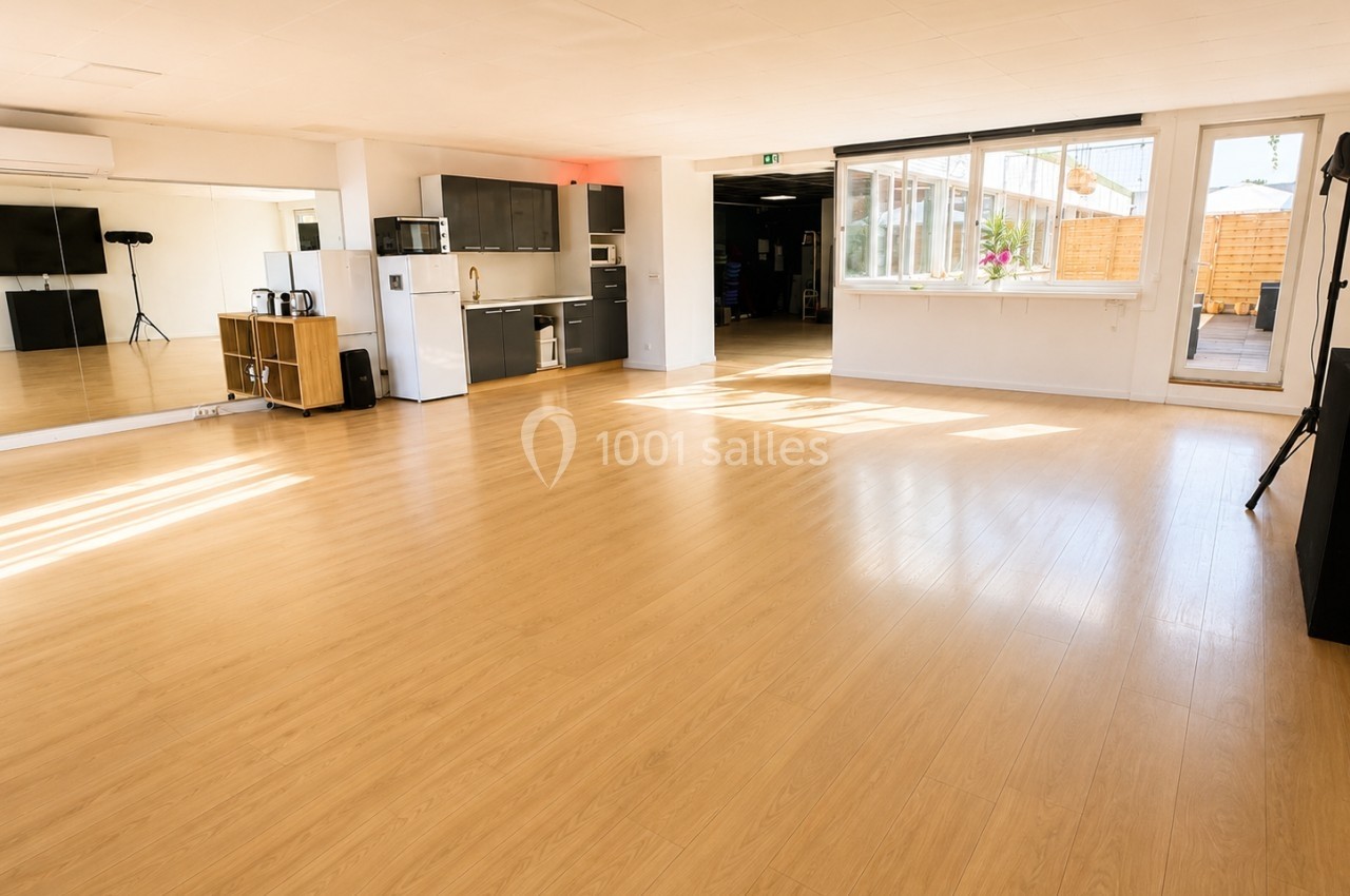 Salle lumineuse avec parquet, miroirs muraux, kitchenette équipée et grandes fenêtres donnant sur une terrasse.