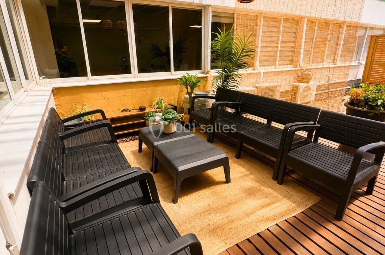 Terrasse aménagée avec des fauteuils noirs, une table basse, des plantes et un tapis en fibre naturelle.