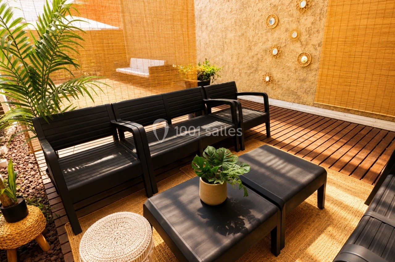 Mobilier de jardin noir sur une terrasse en bois, décorée de plantes, tapis en fibres naturelles et murs beige ornés.