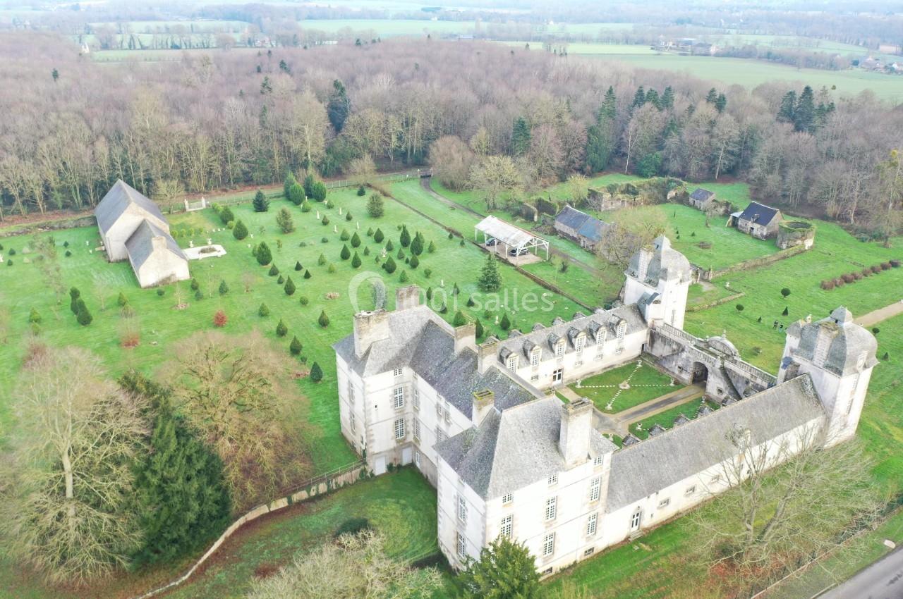Vue aérienne d'un château entouré de jardins, d'arbres et de bâtiments annexes au milieu d'une campagne boisée.