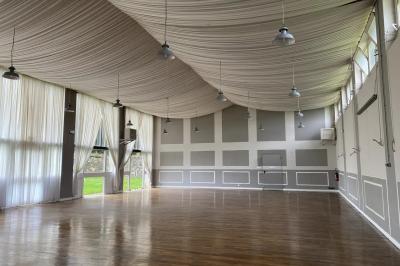 Grande salle vide avec parquet en bois, murs gris et blancs, plafond drapé de tissu beige et grandes fenêtres laissant…