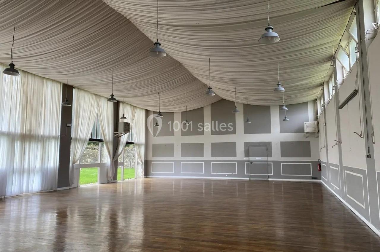 Grande salle vide avec parquet en bois, murs gris et blancs, plafond drapé de tissu beige et grandes fenêtres laissant…