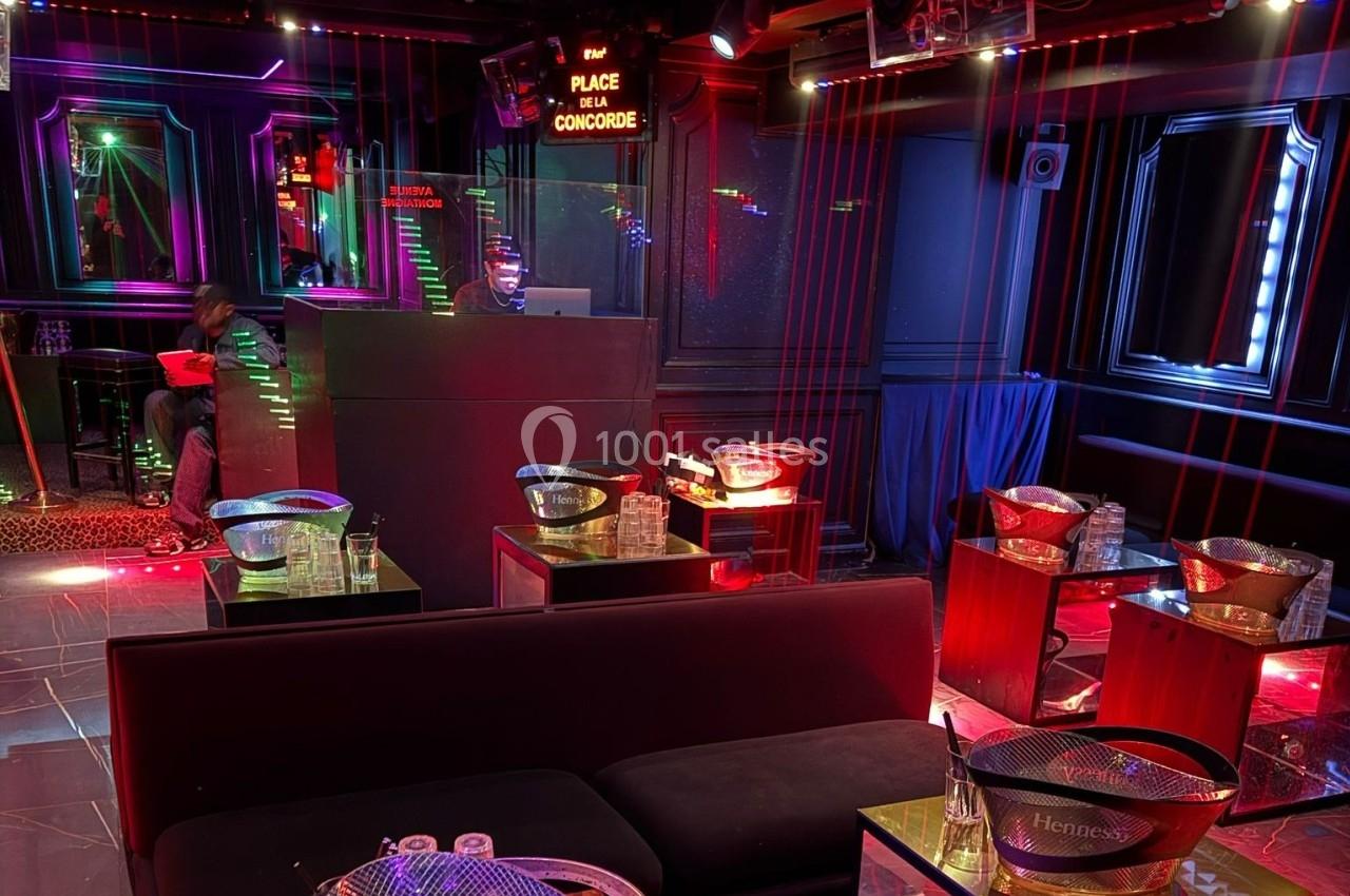 Salle de boîte de nuit avec éclairage néon, tables basses équipées de seaux à glace et banquettes noires.