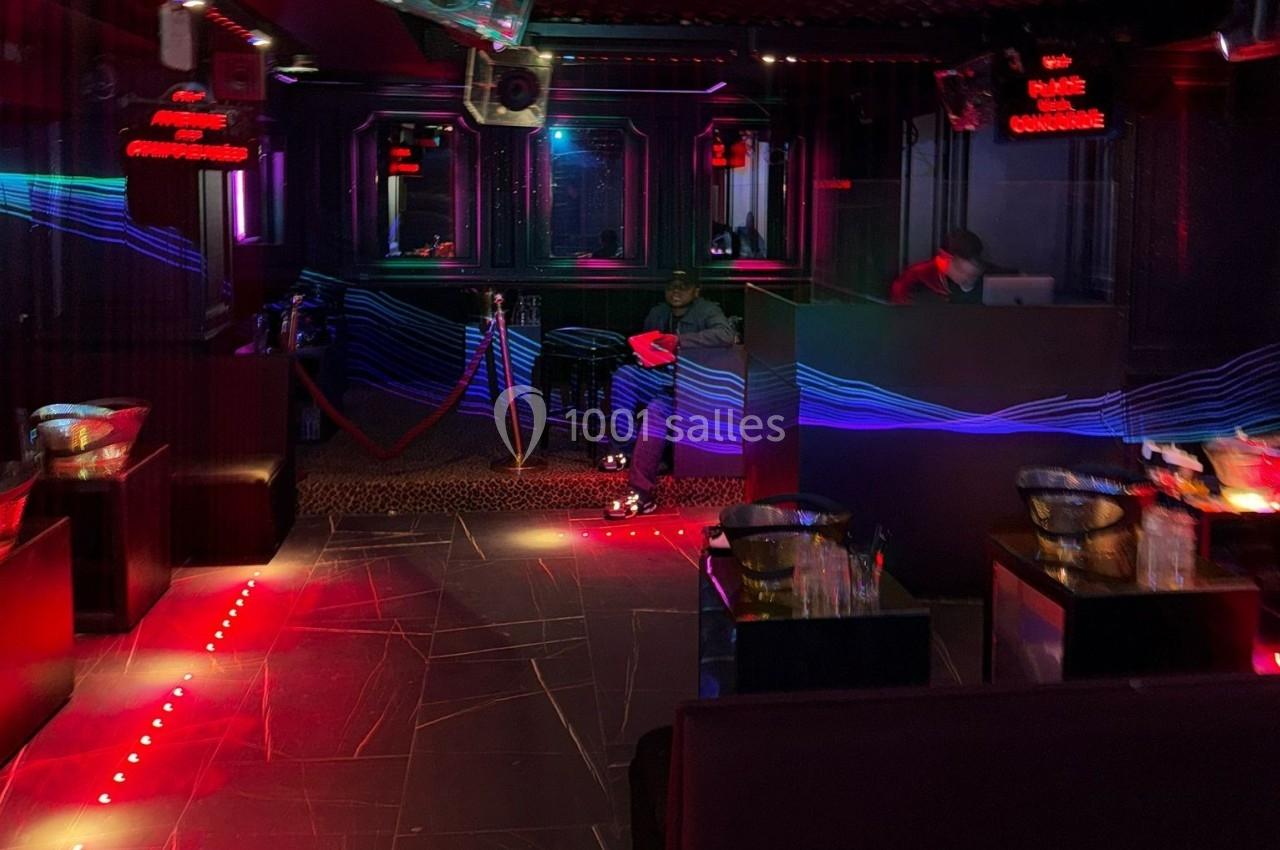 Salle de nuit sombre avec éclairages rouges et bleus, un comptoir à droite et des tables disposées autour.
