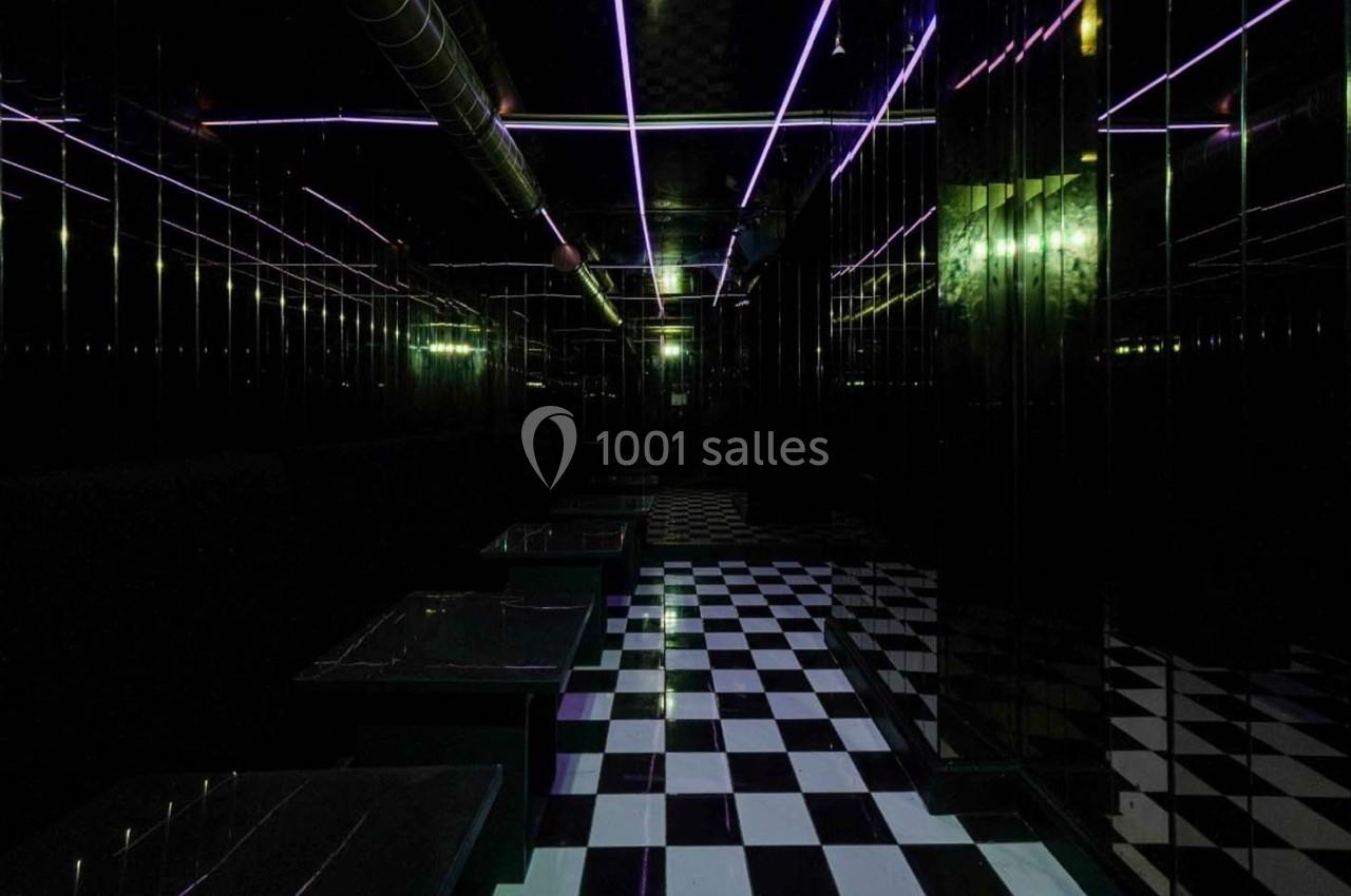 Salle sombre avec sol à damier noir et blanc, murs réfléchissants et éclairage néon violet et vert.