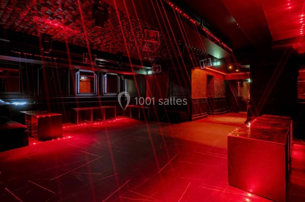 Salle de club vide avec éclairage rouge, murs sombres, plafond texturé et mobilier minimaliste.