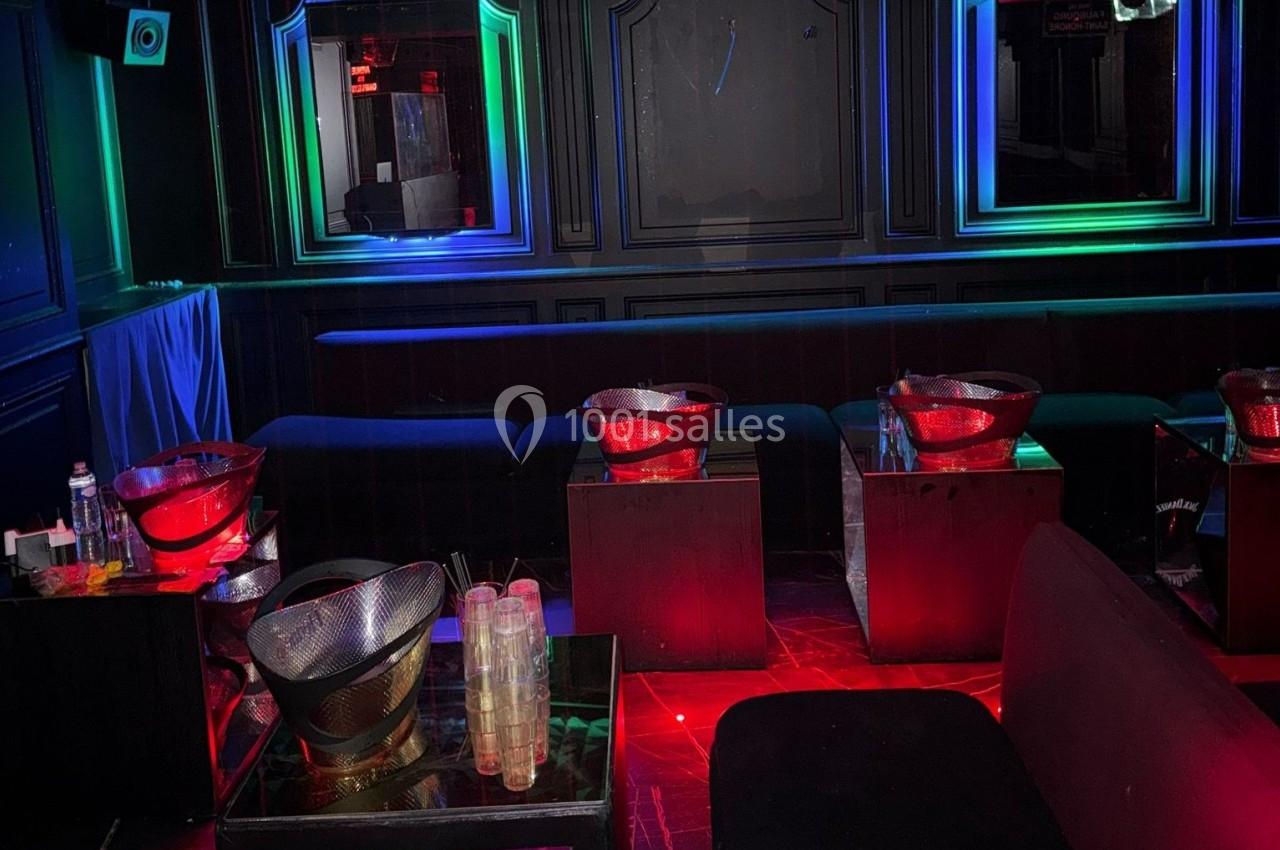 Salle de réception sombre avec banquettes, tables basses, lumières colorées et seaux à glace rouges.