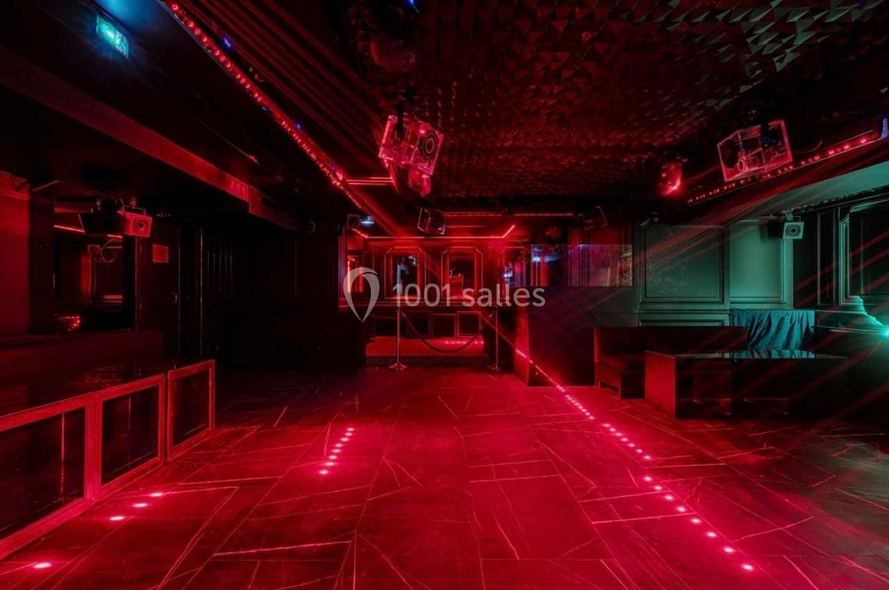 Salle de club vide avec éclairage rouge, murs sombres, sol texturé et équipements audio visibles.