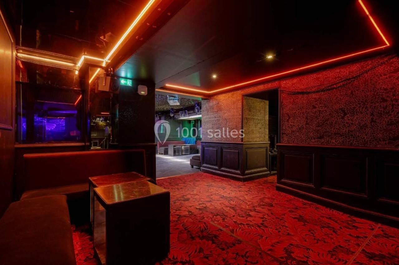 Salle intérieure avec banquettes, table basse, éclairage rouge et décorations sombres, donnant sur un espace ouvert.