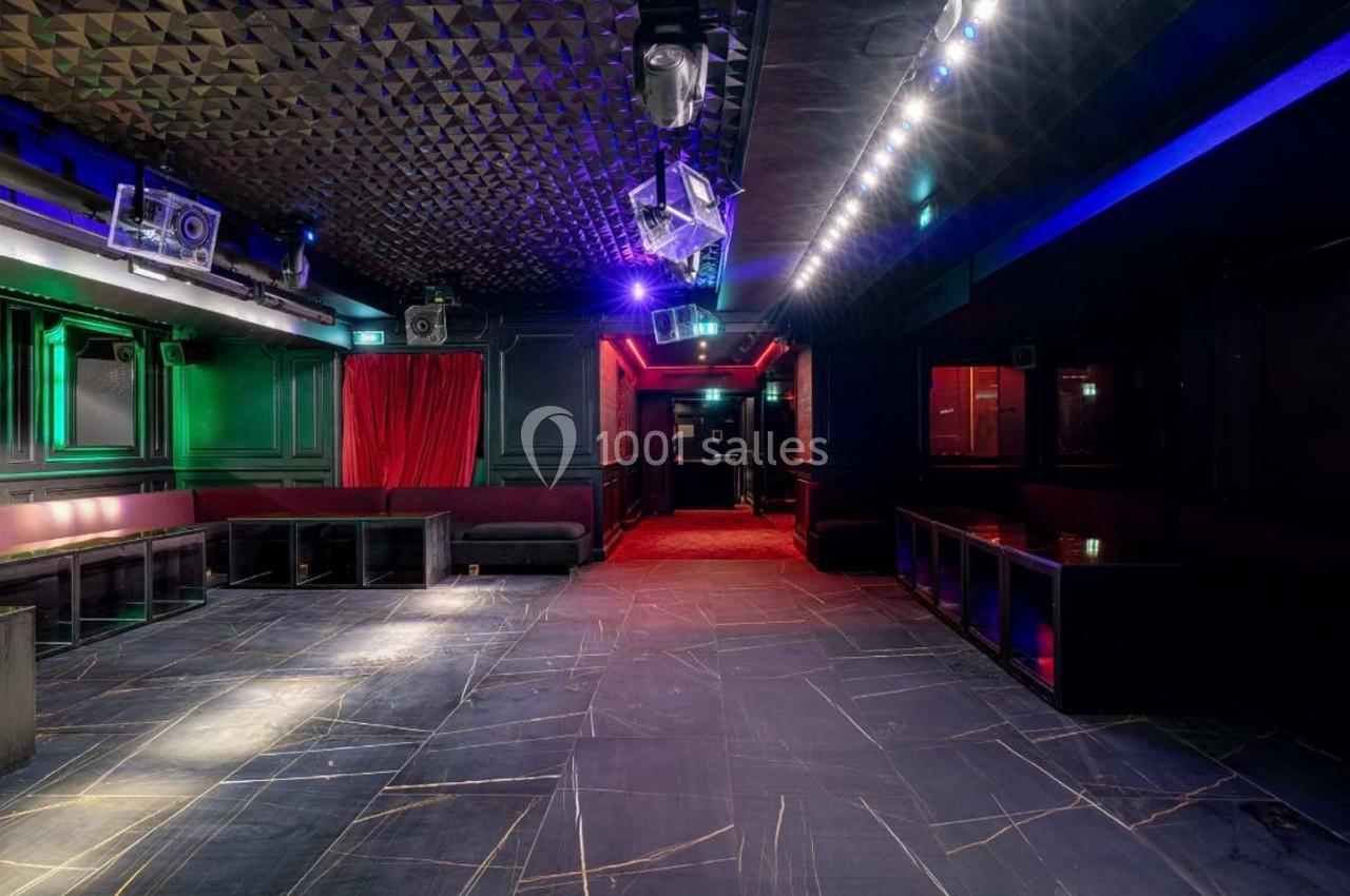 Salle de club vide avec éclairage tamisé, murs sombres, banquettes et sol en carrelage noir.