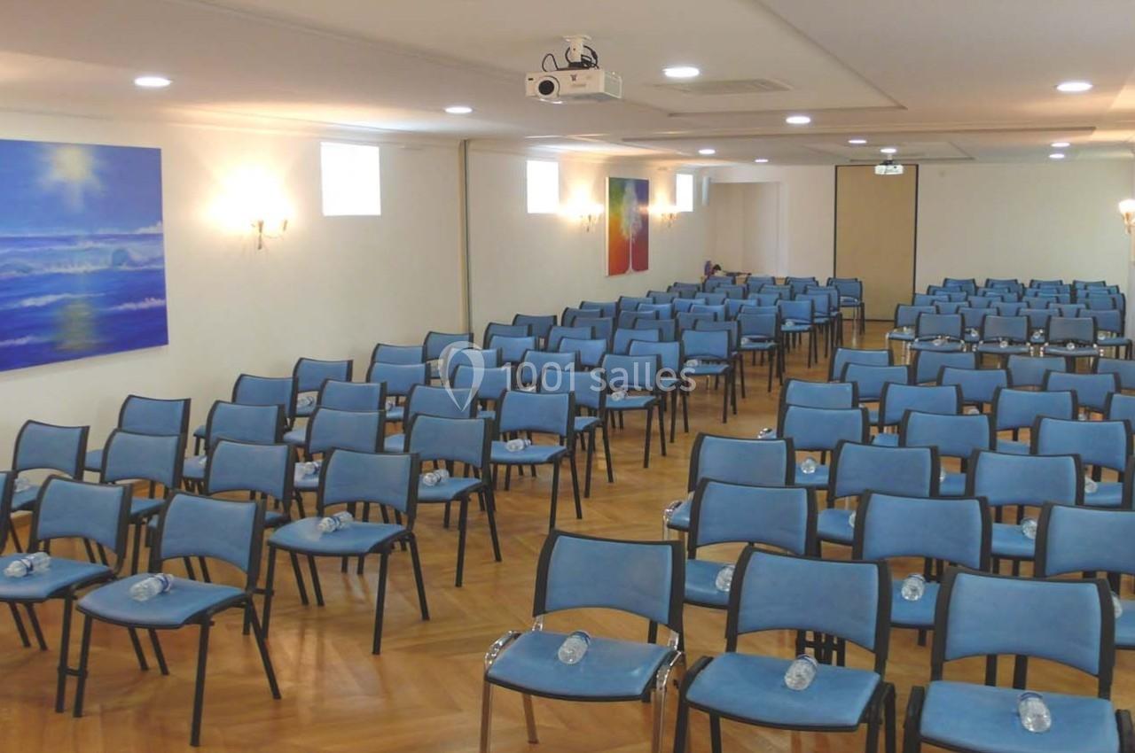 Salle de conférence lumineuse avec des rangées de chaises bleues alignées et des bouteilles d'eau posées sur chaque siège.